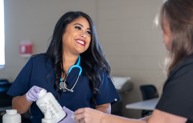 Two healthcare professionals in scrubs are smiling and engaging in conversation, one holding a medication bottle in a clinical setting.